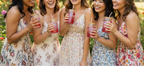 Five women in floral dresses holding red drinks with straws outdoors.