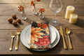 Dining table setting with floral napkin, plates, cutlery, and candles on a wooden surface.