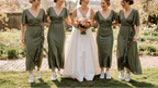 Bride in a white dress with floral bouquet surrounded by four women in green dresses outdoors.