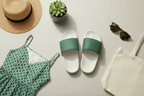 Green and white slide sandals with a dress, hat, plant, sunglasses, and tote bag on a light surface.
